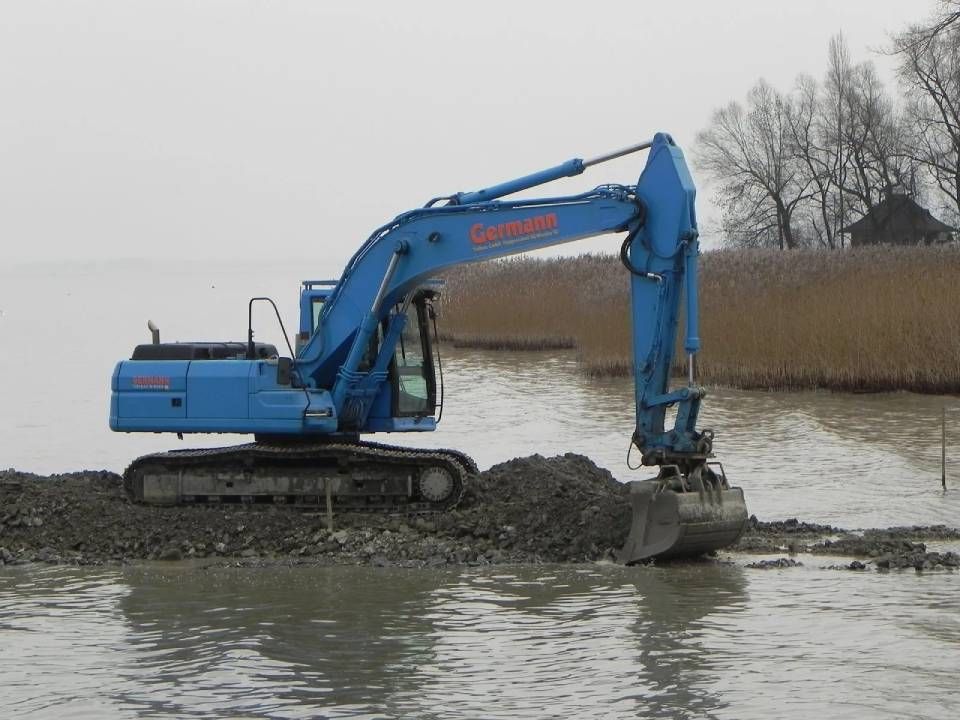 Blauer Bagger im Wasser, der in Ufernähe Treibgut aufnimmt, bewölkter Tag.