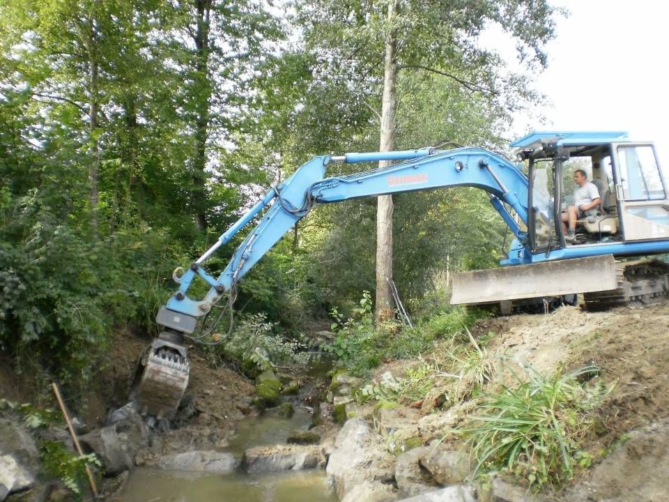 Ein blauer Bagger ist in der Nähe eines Bachs im Einsatz, eine Person befindet sich in der Kabine und arbeitet in einem Waldgebiet.