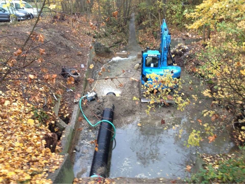 Ein blauer Bagger arbeitet an einem grossen schwarzen Rohr in einem schlammigen, wassergefüllten Graben in einem Waldgebiet.