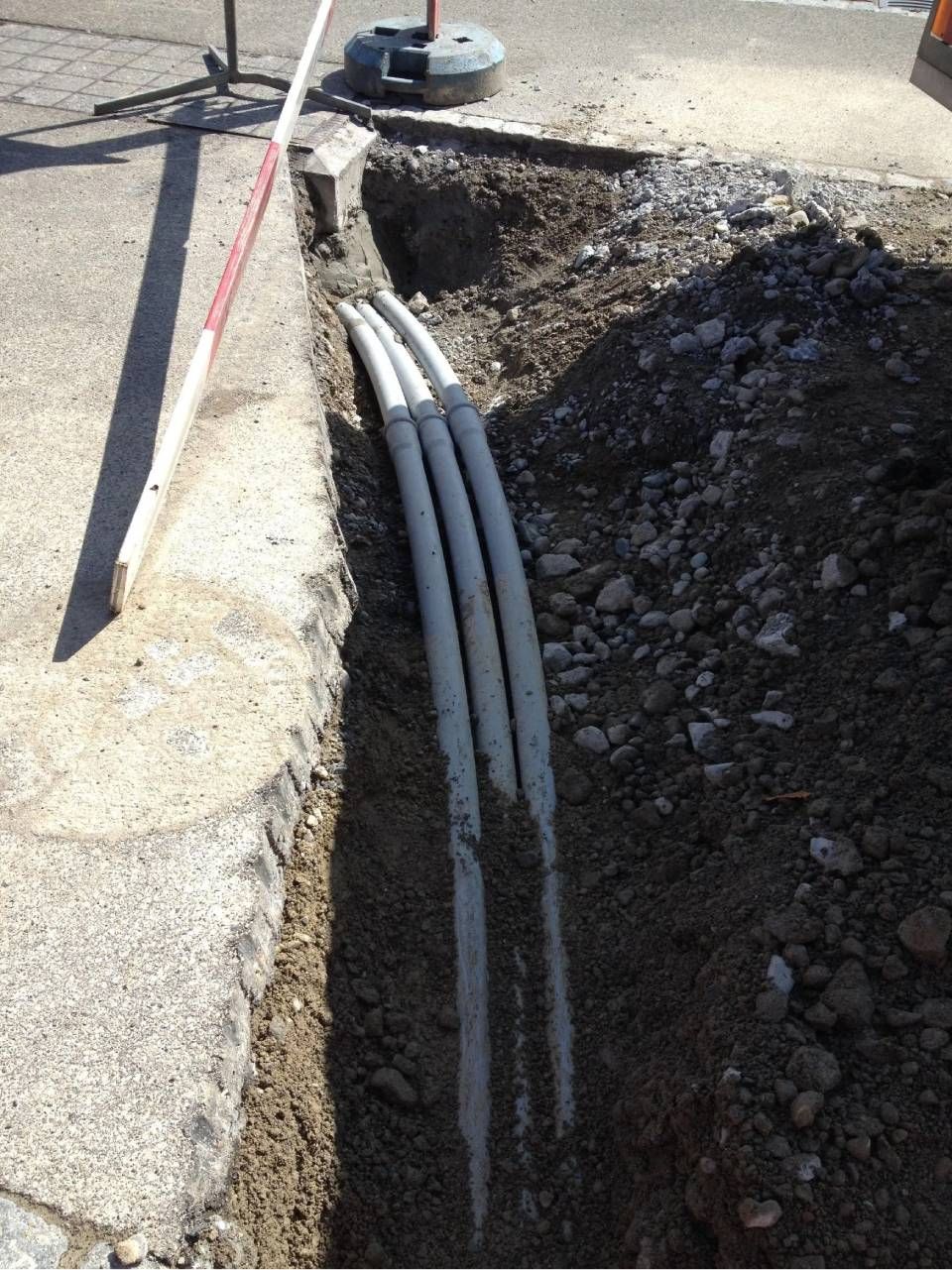 Drei graue Leitungen in einem offenen Graben neben einer Betonbordsteinkante. Ein weiss-roter Mast ist zu sehen.