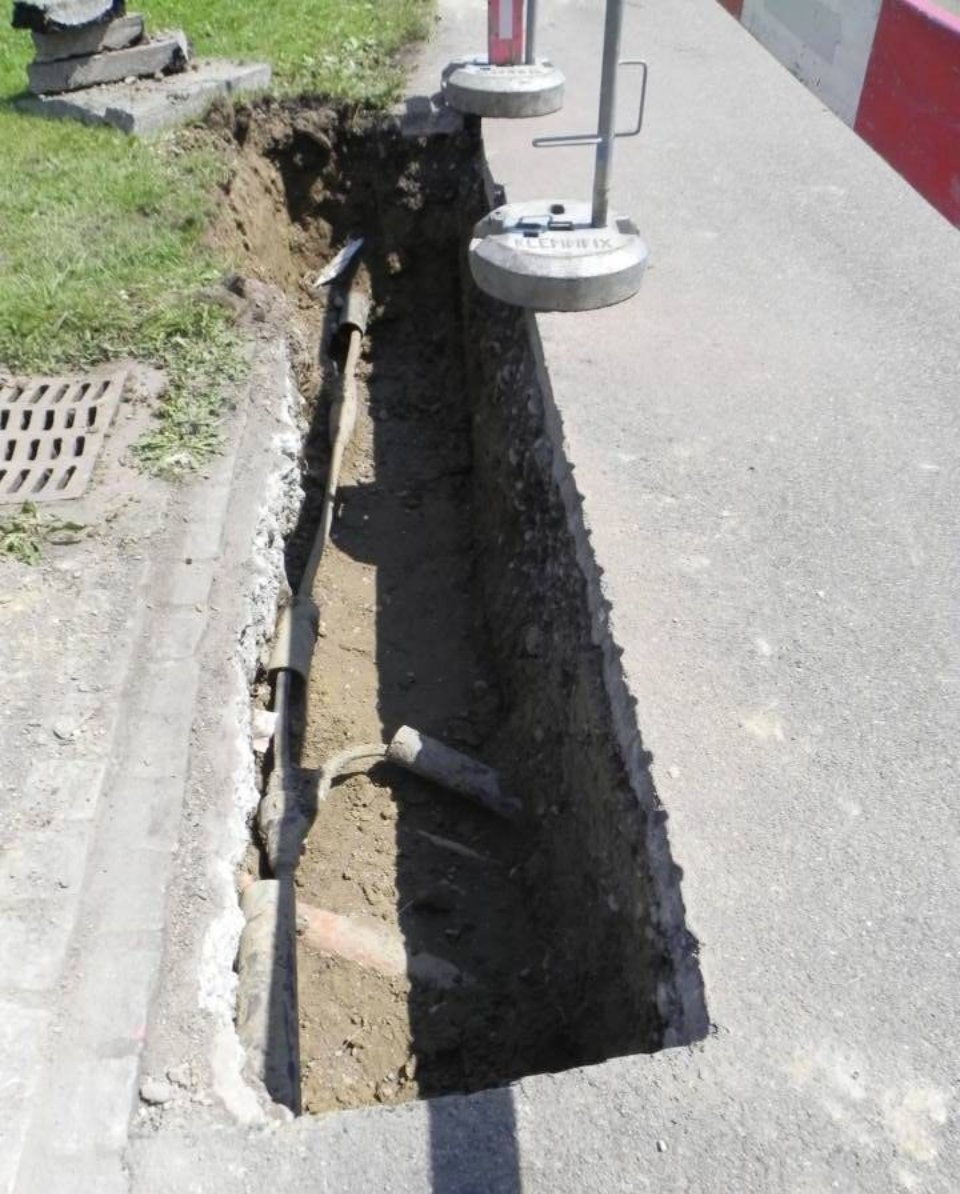 Offener Graben im Asphalt neben einem Bordstein und einer Rasenfläche. Betonblöcke und Rohre sind im Graben sichtbar.