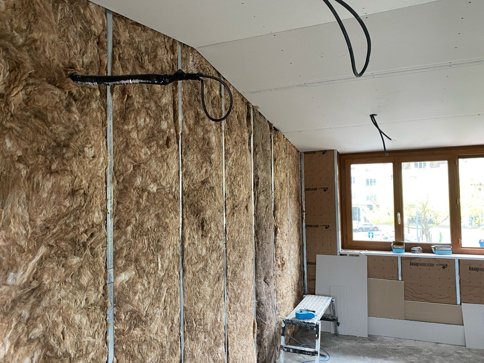 Pièce inachevée avec isolation apparente sur les murs, câbles électriques au plafond et fenêtres au fond.