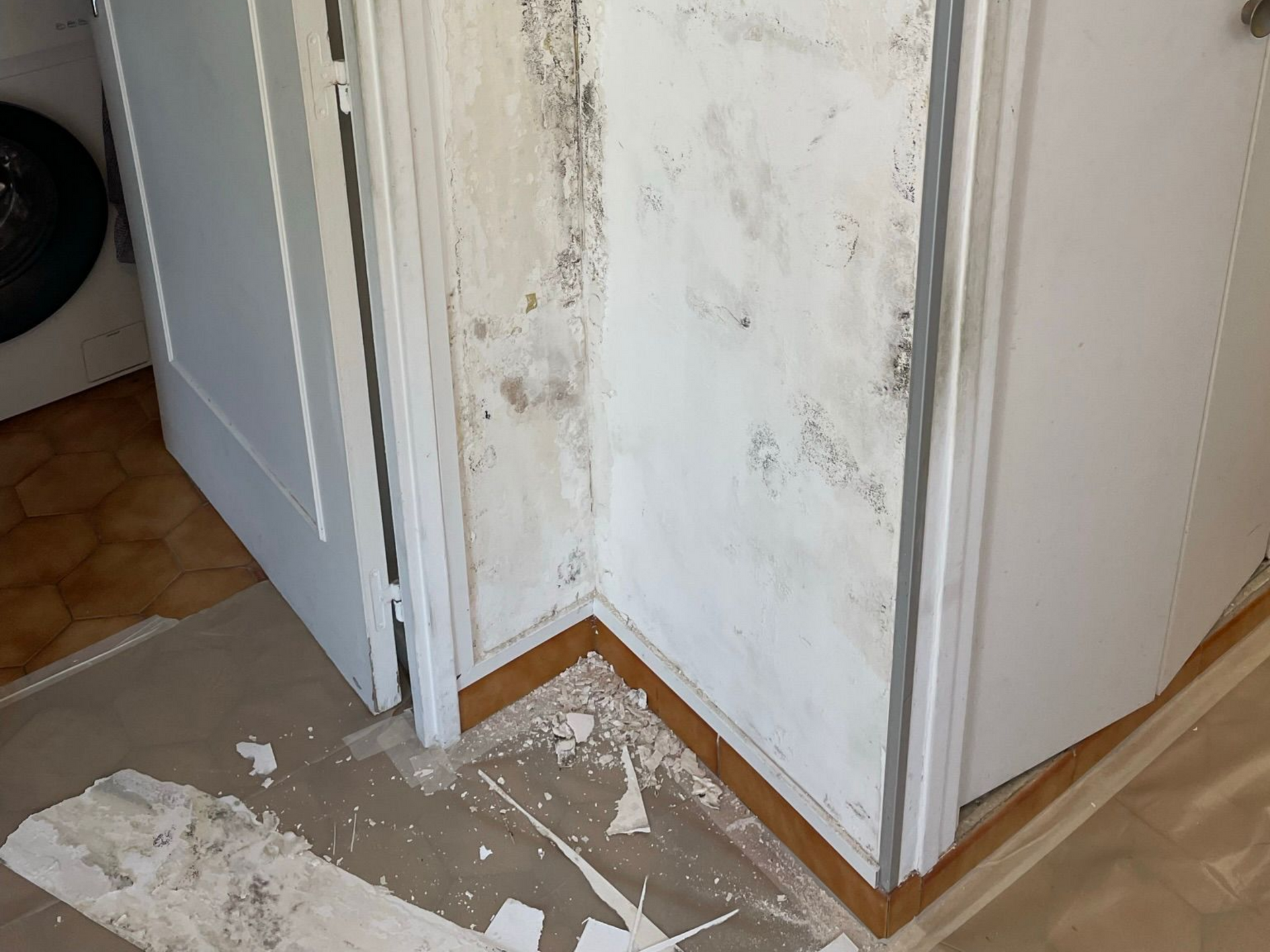 Mur et cadre de porte endommagés, peinture écaillée et débris au sol dans un couloir étroit.