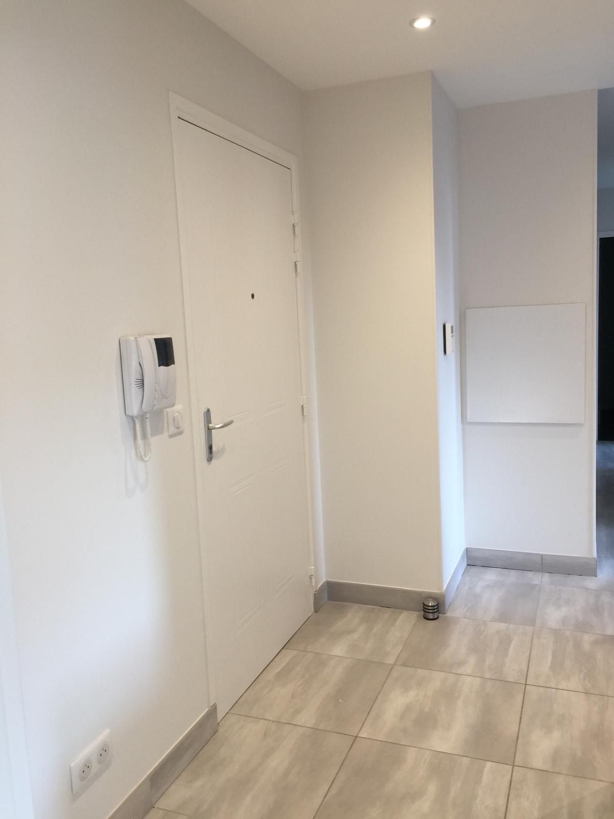 Couloir moderne avec porte blanche, interphone mural, carrelage clair au sol et spots encastrés au plafond.
