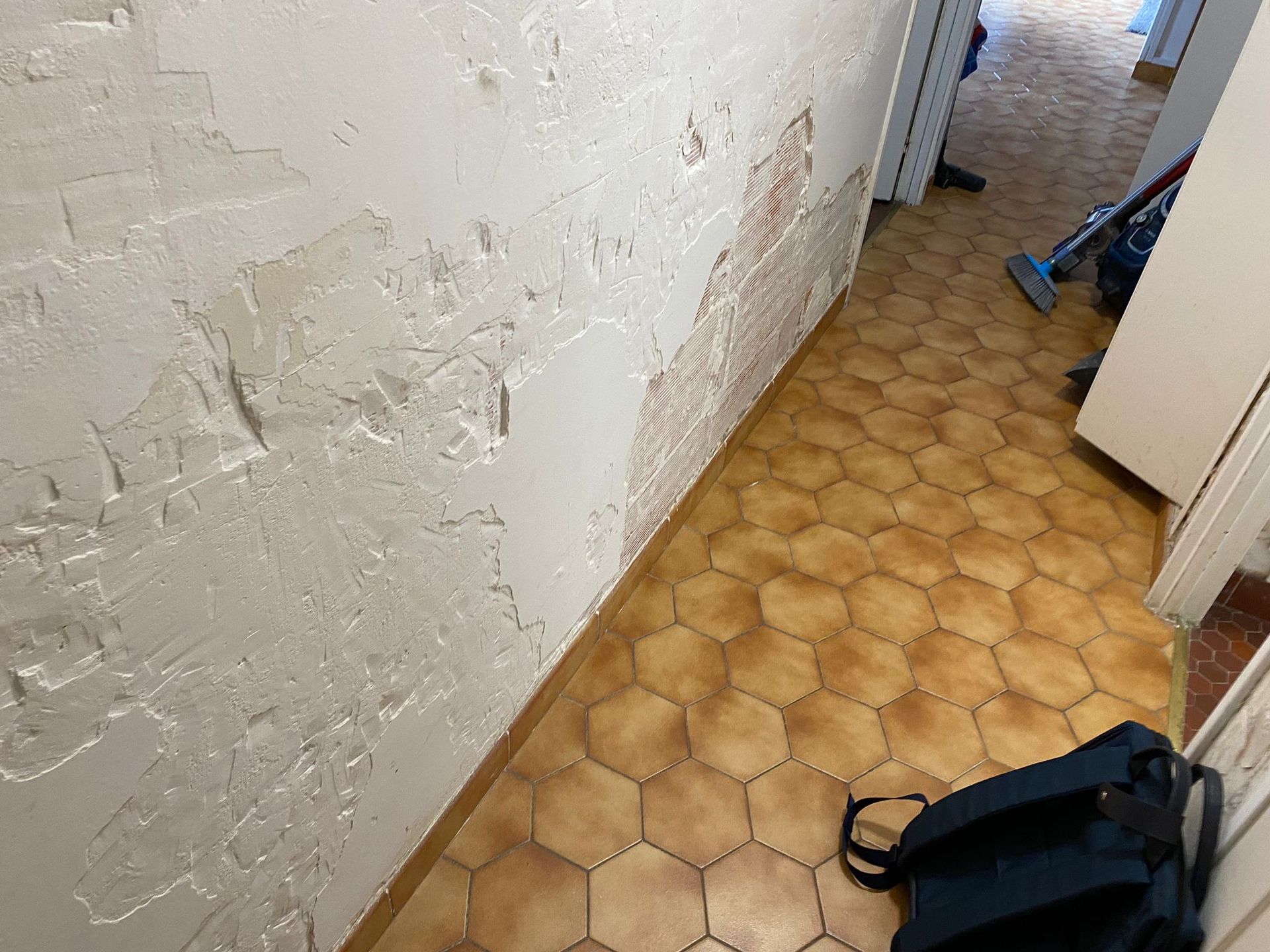 Couloir étroit avec un mur blanc écaillé, un sol carrelé beige et un sac noir posé au sol près d'une porte.