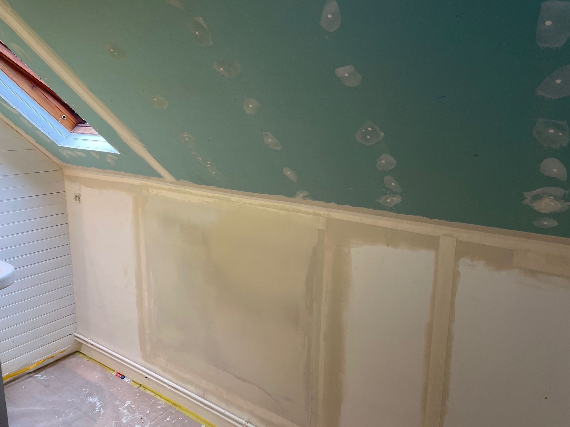 Pièce mansardée partiellement aménagée avec plafond mansardé, murs bruts avant mise en peinture et petite fenêtre de toit.