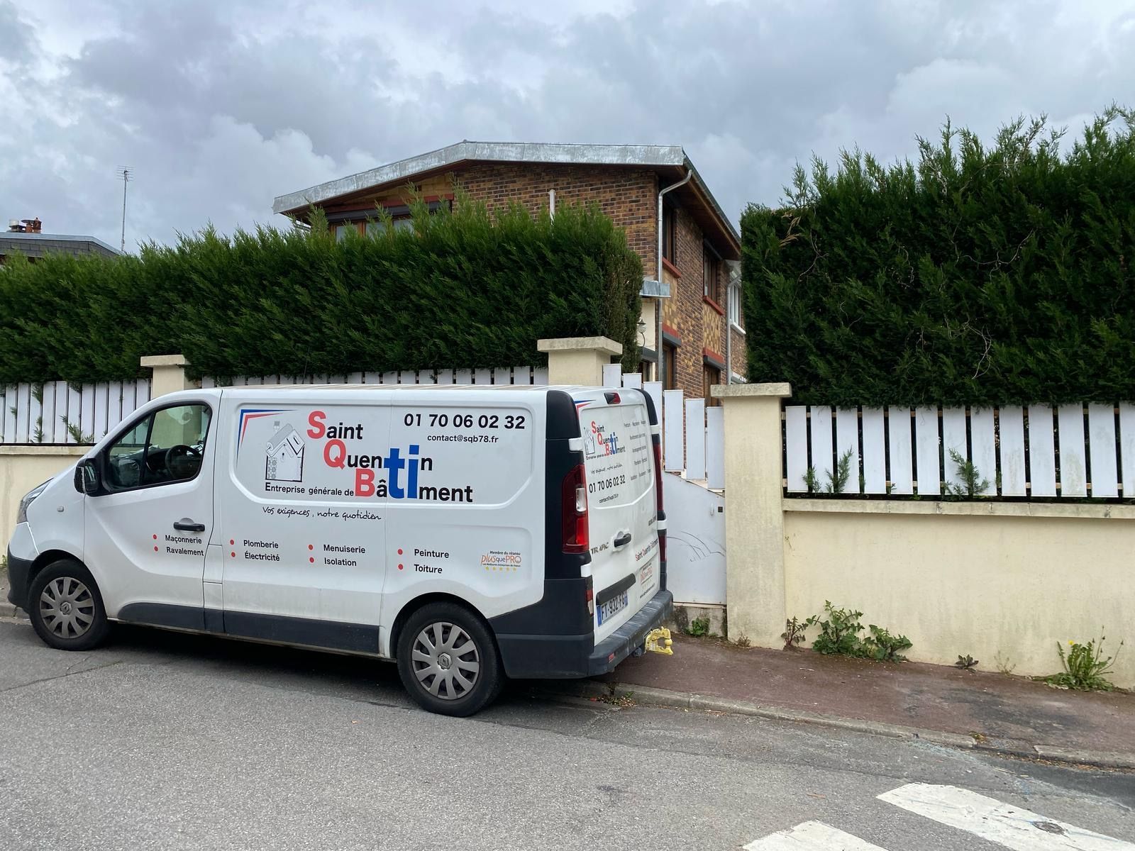 Une camionnette d'artisan est garée devant une maison en briques et des haies..