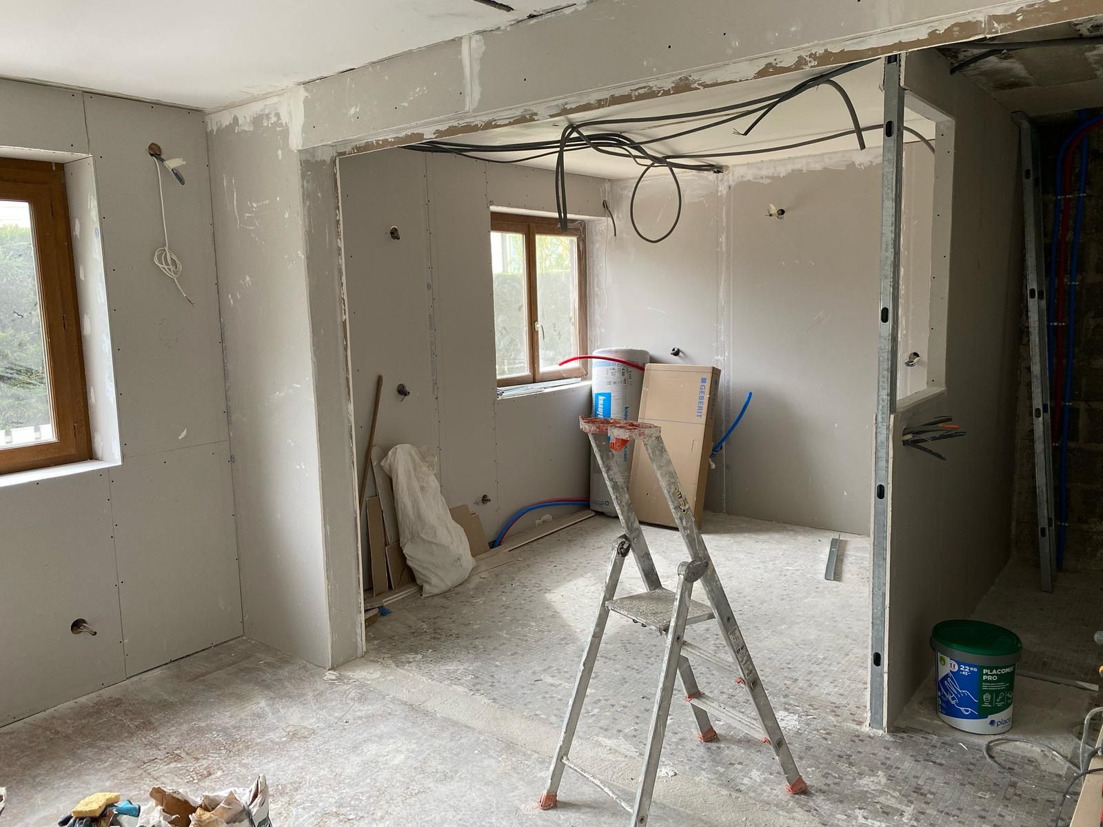 Pièce partiellement rénovée avec cloisons sèches, escabeau, câbles électriques apparents et outils de chantier au sol.