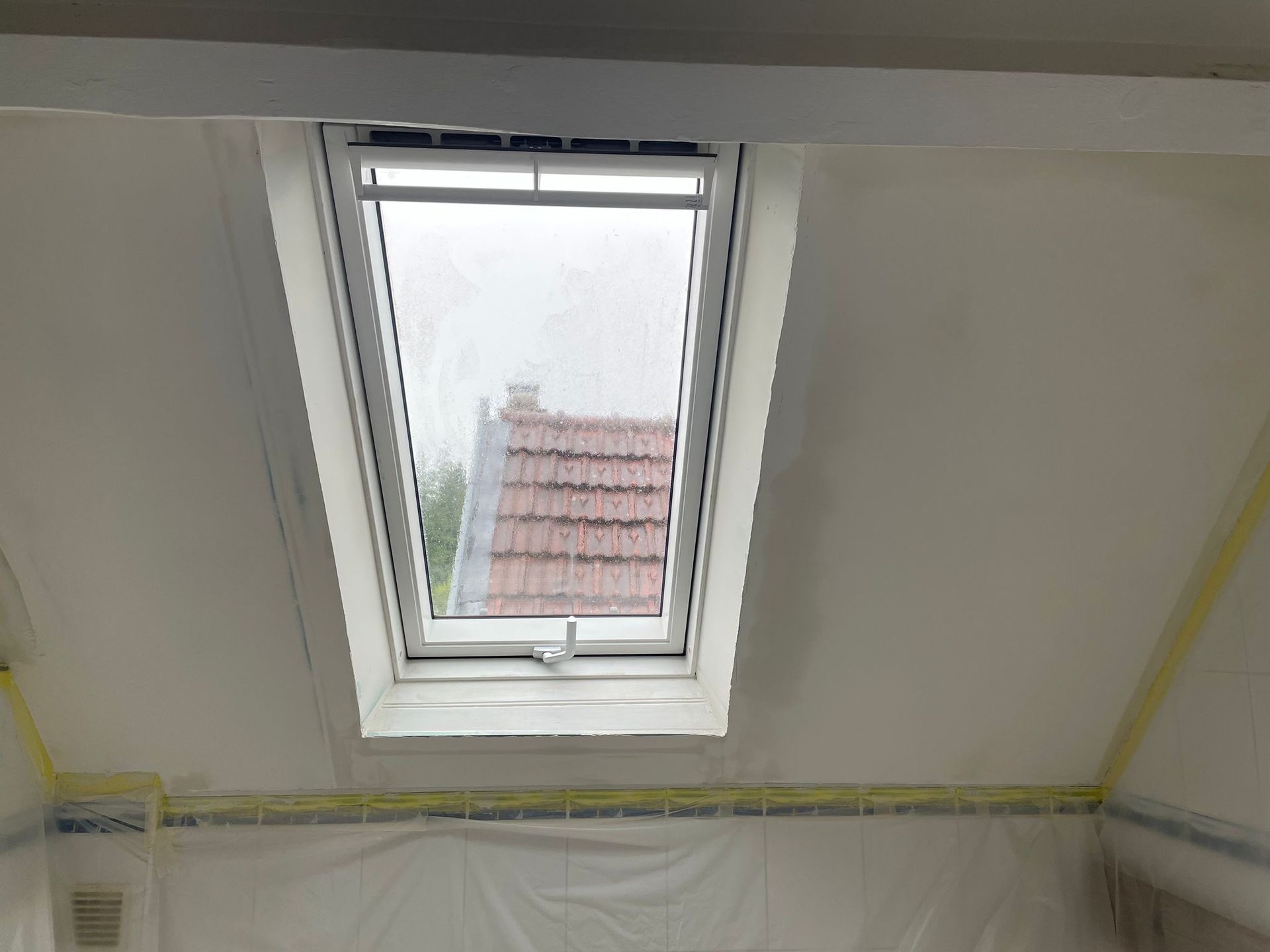 Fenêtre de toit dans une pièce blanche, avec du plastique de protection et du ruban de masquage le long des murs.