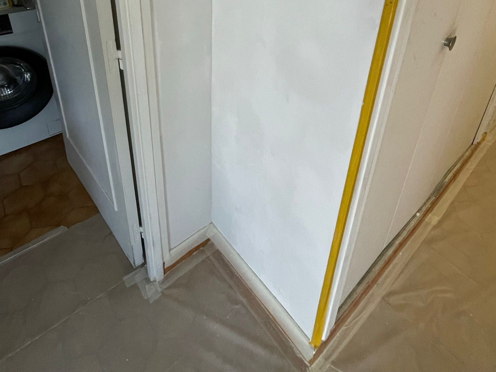 Coin d'une pièce avec une porte blanche, un mur rénové aux bords jaunes et un sol protégé par une bâche grise.