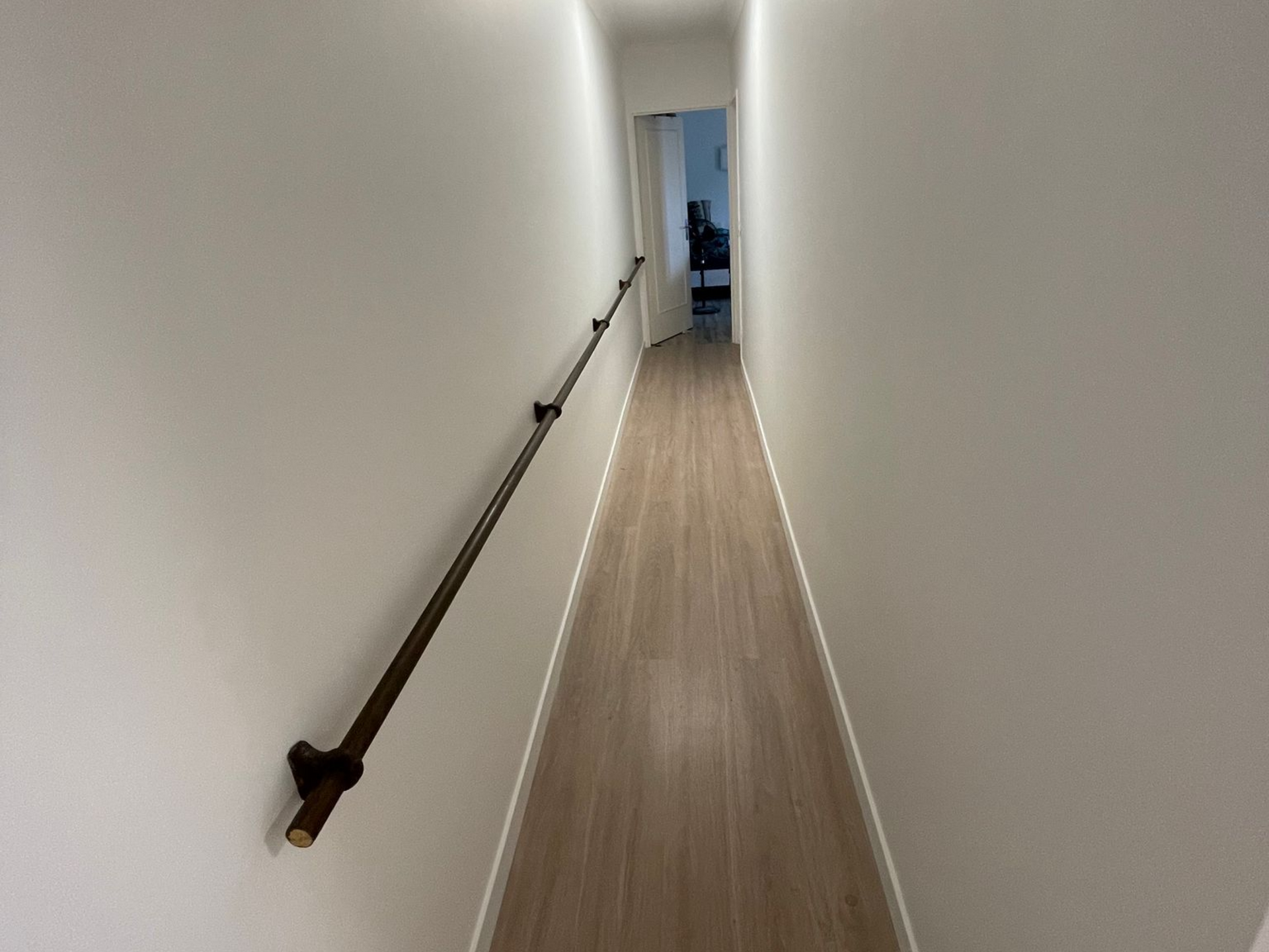 Couloir étroit aux murs blancs, au sol en bois et doté d'une rampe sur le côté gauche.
