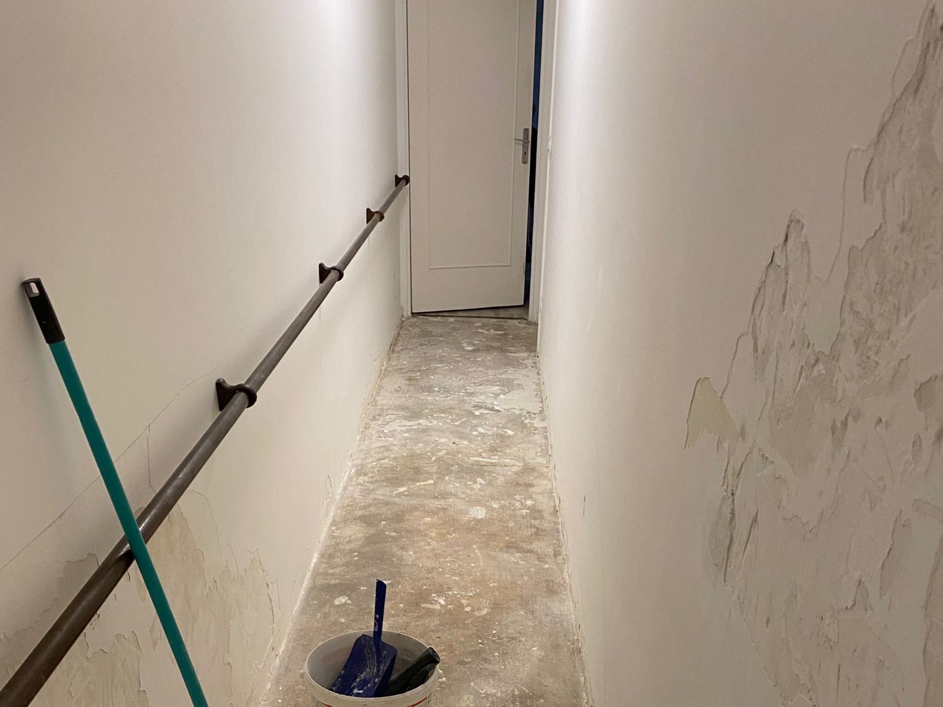 Couloir étroit et inachevé avec une serpillière, un seau et une porte entrouverte au fond.