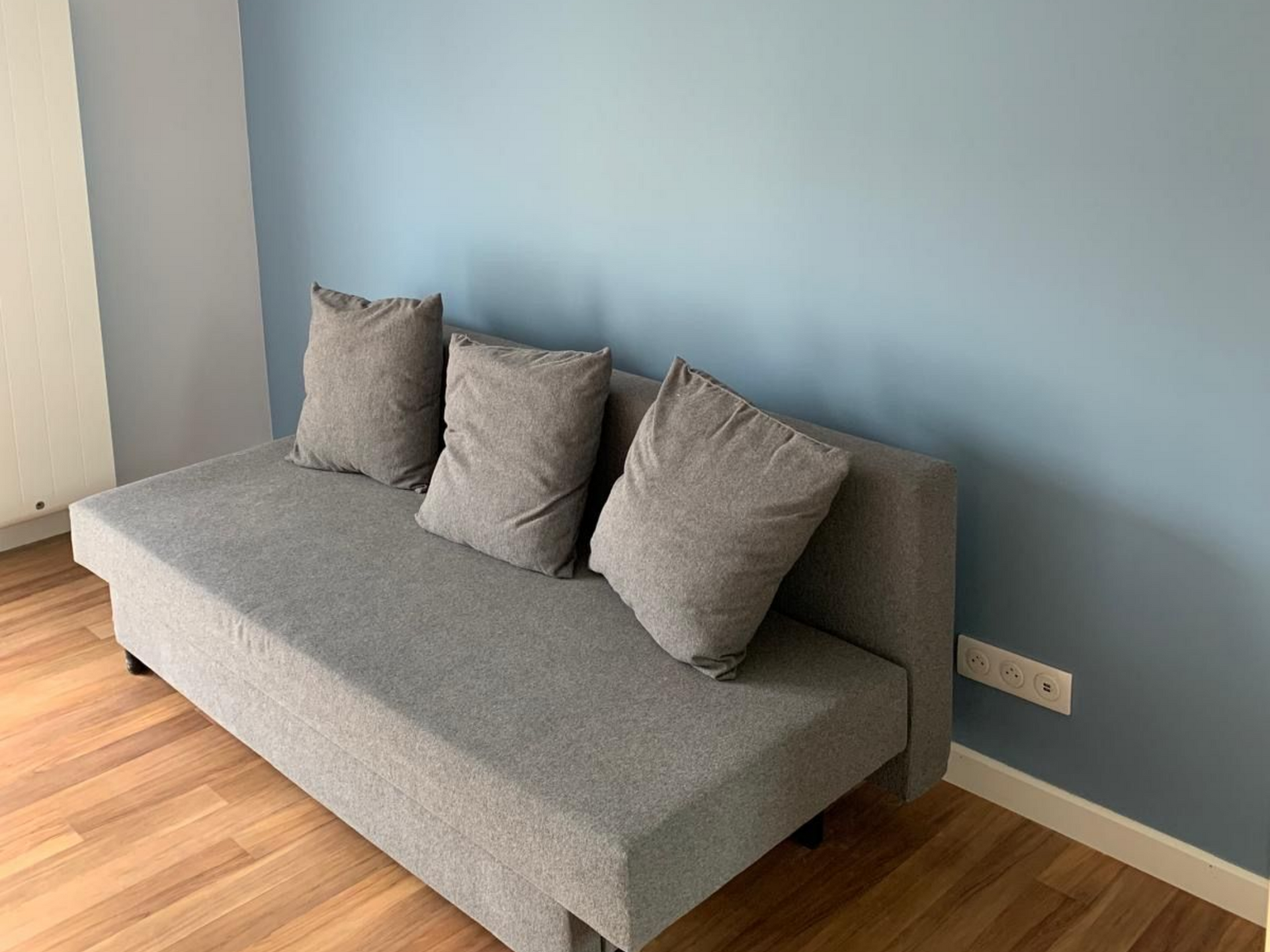 Canapé gris avec trois coussins contre un mur bleu clair dans une pièce lumineuse.