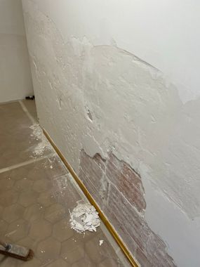 Mur intérieur endommagé, peinture blanche écaillée et plâtre apparent près d'un sol carrelé.