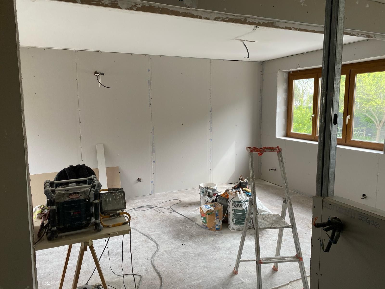 Pièce partiellement rénovée avec la pose de plaques de plâtre, escabeau, outils et une fenêtre laissant entrer la lumière du jour.