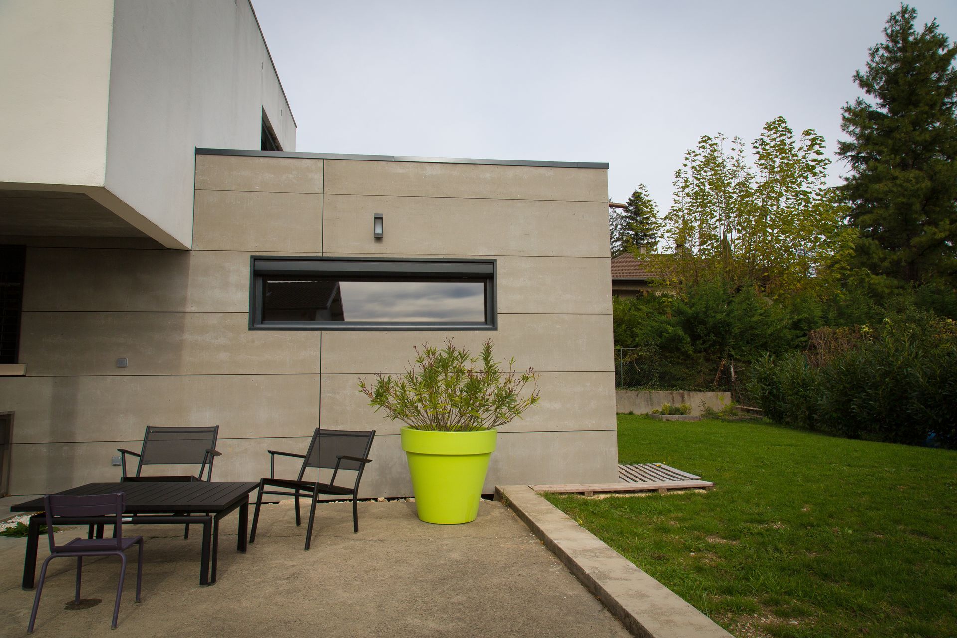 Terrasse moderne avec chaises d'extérieur noires, jardinière vert vif et mur de maison beige percé d'une fenêtre étroite.
