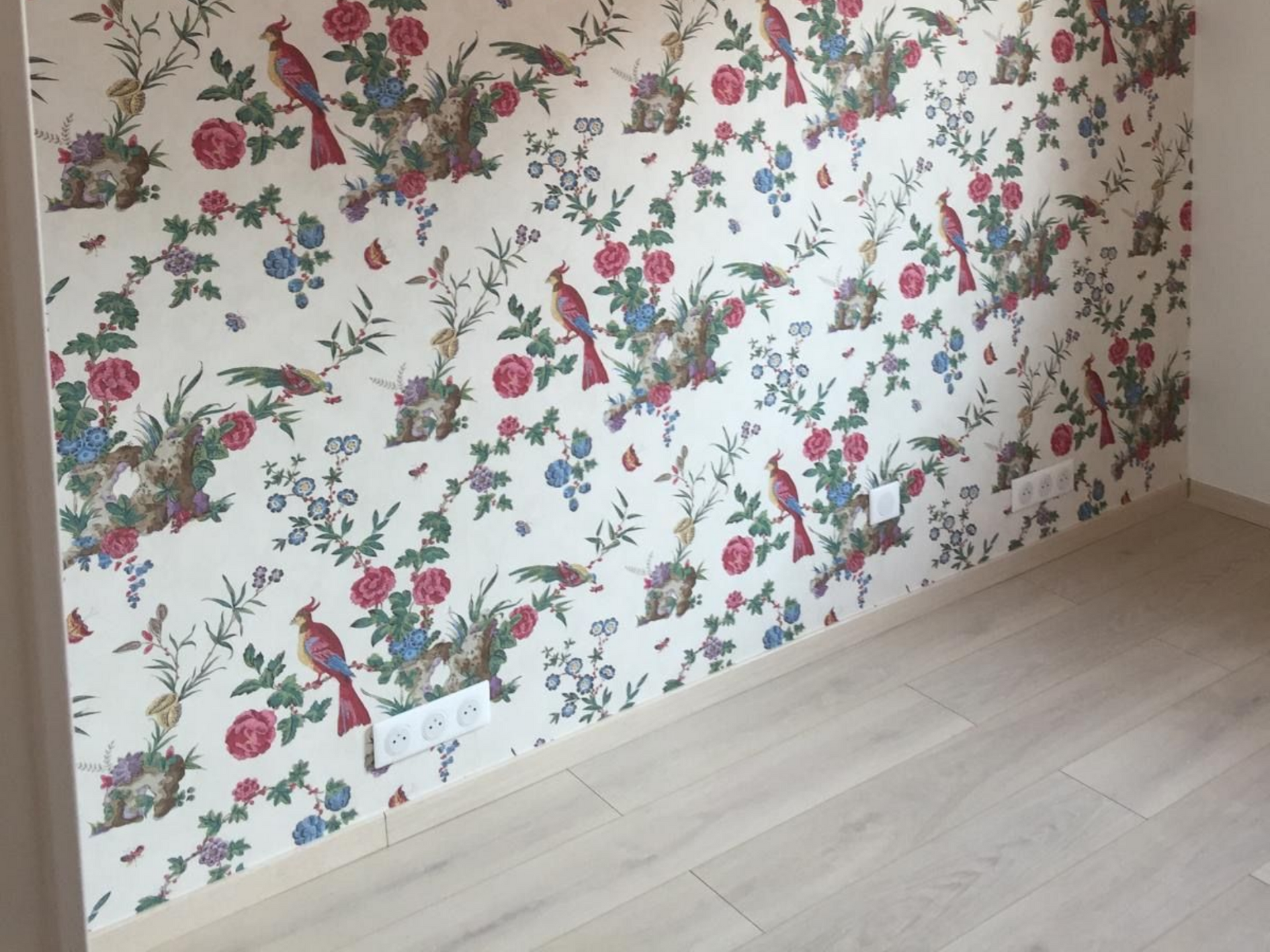 Mur tapissé de papier peint à motifs floraux avec des prises électriques blanches au-dessus d'un parquet clair.