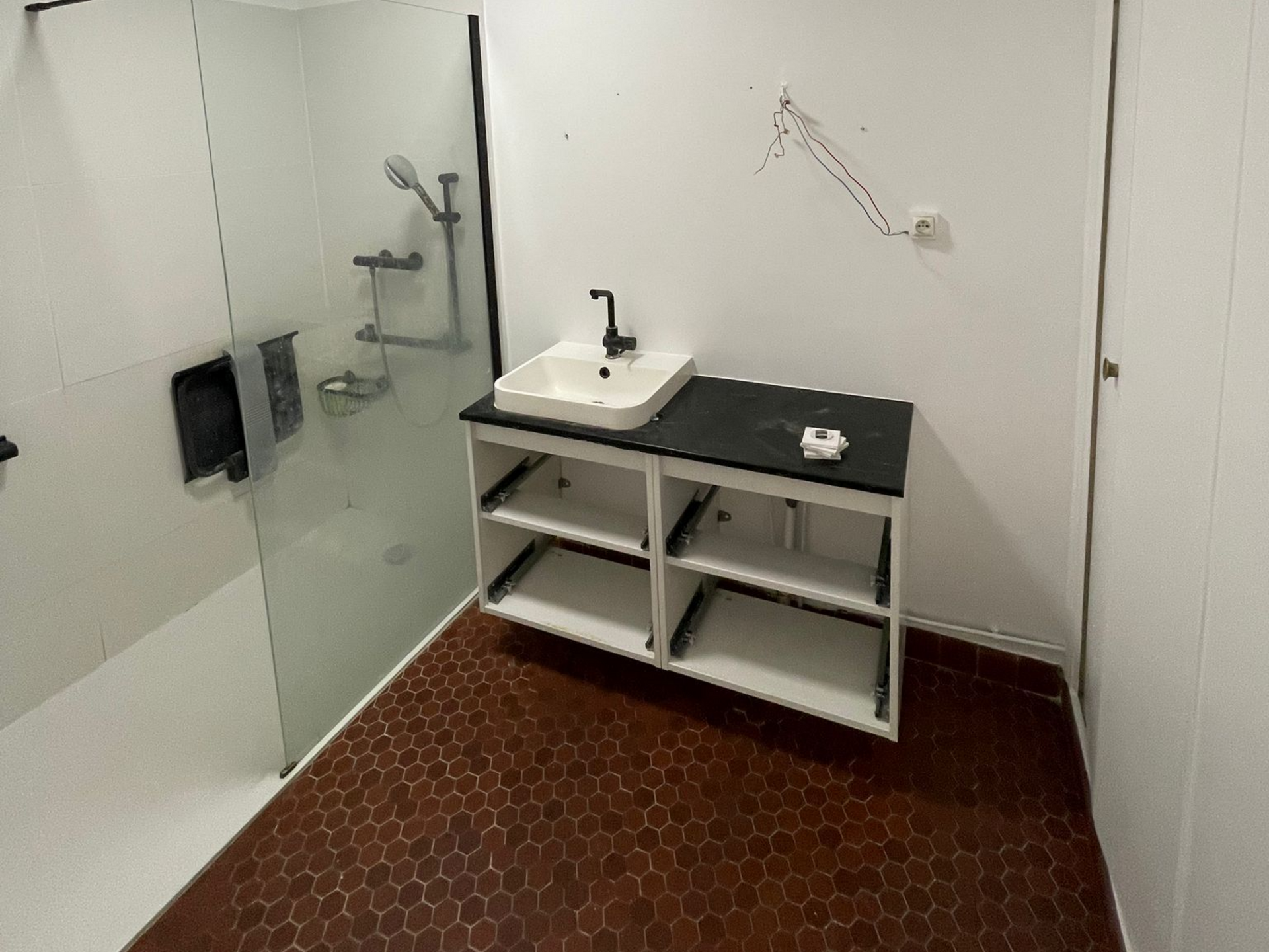 Salle de bains moderne avec douche, lavabo blanc et étagères foncées sur murs blancs et sol carrelé de carreaux hexagonaux.