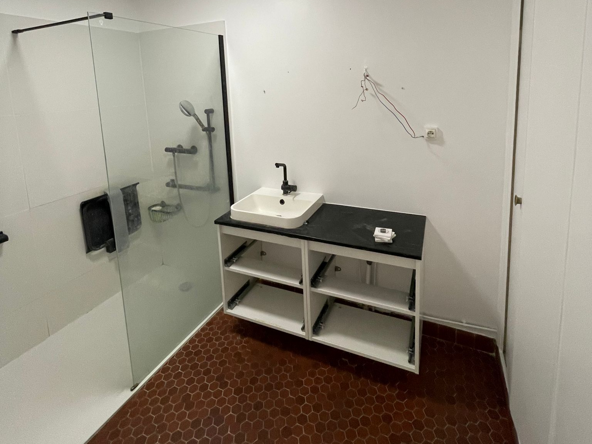 Salle de bains moderne avec douche, lavabo blanc, plan de travail noir et carrelage hexagonal au sol.