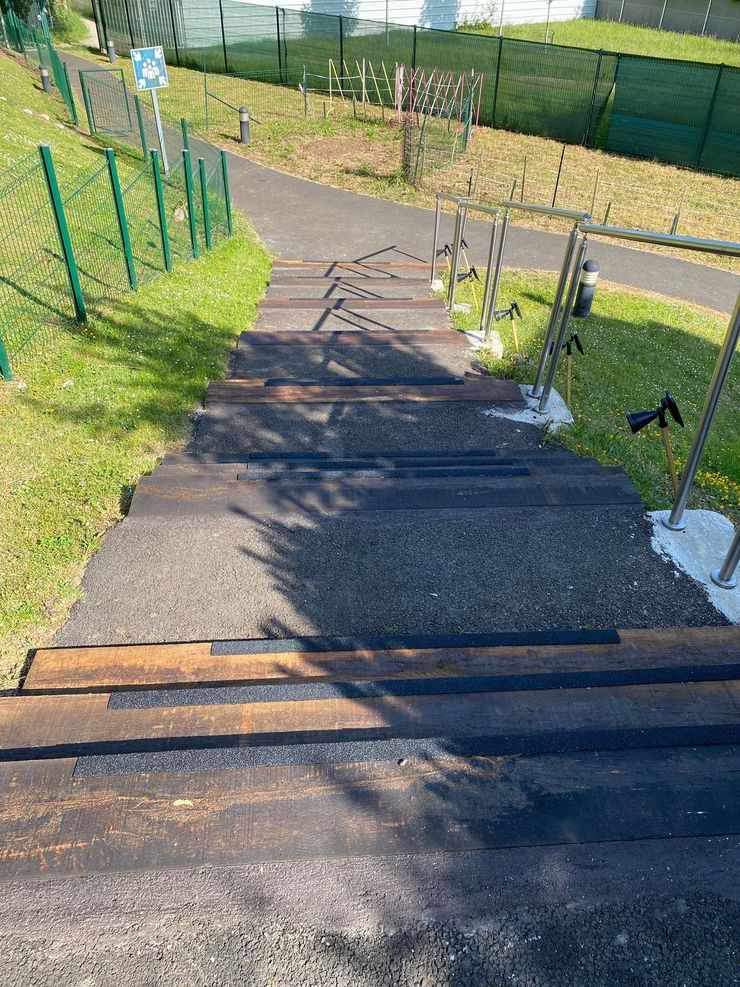 Escalier extérieur avec marches aux bords en bois donnant sur un chemin en gravier, bordé d'une clôture verte et de pelouse ensoleillée.