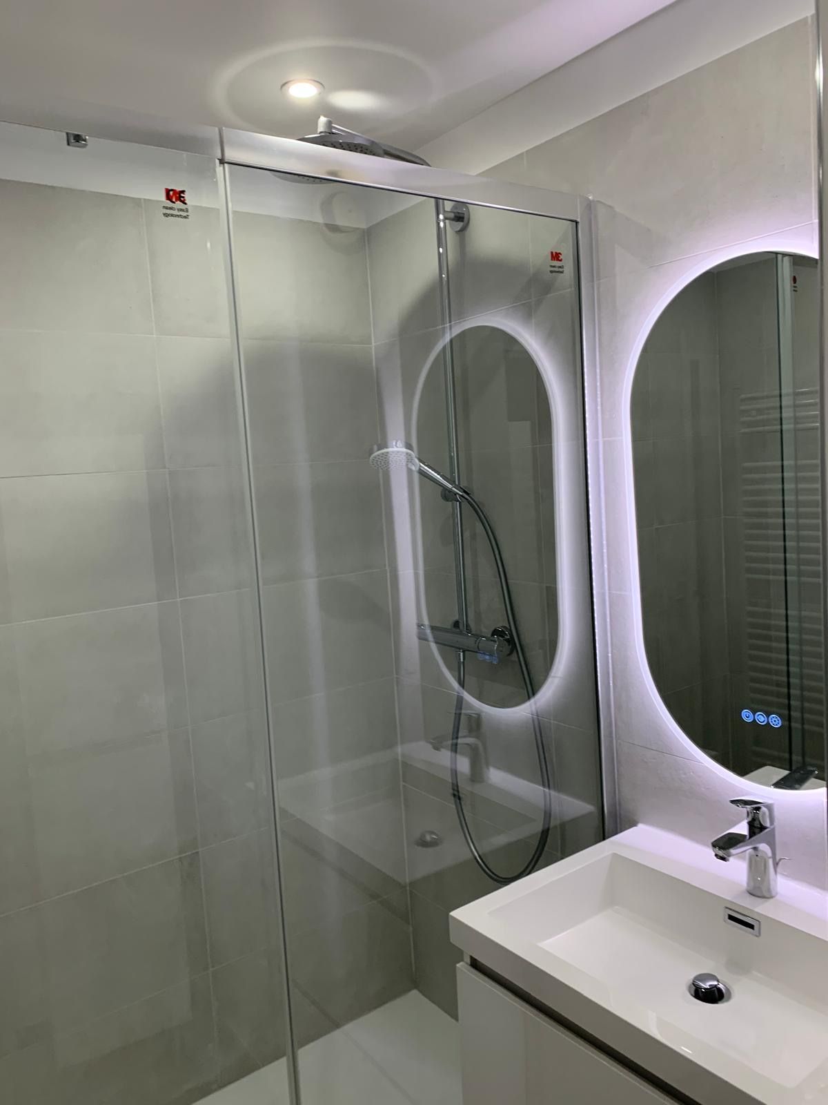 Salle de bains moderne avec douche, miroir ovale rétro éclairé et lavabo blanc.