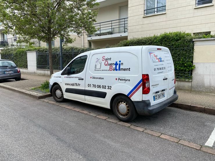 Une camionnette d'artisan est garée dans une rue résidentielle, à côté d'un immeuble et d'arbres.