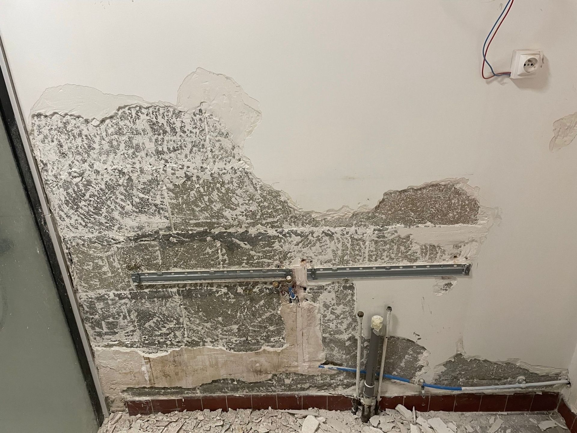 Mur intérieur endommagé avec peinture écaillée, plâtre apparent et tuyaux de plomberie près du sol.