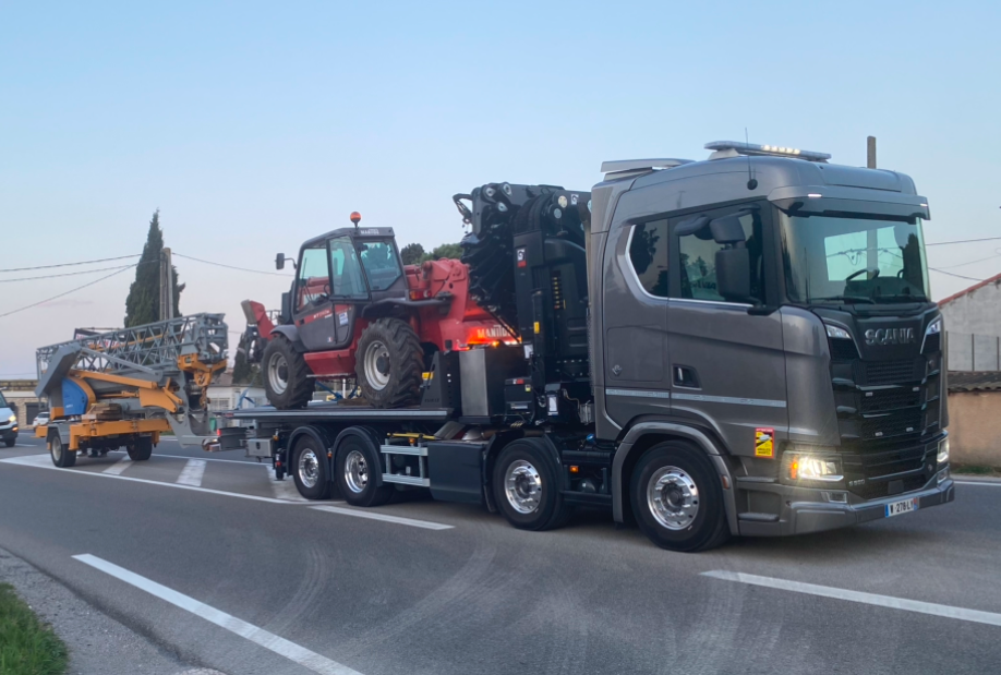 Un semi-remorque gris transporte sur la route des engins lourds, dont un tracteur rouge et une machine jaune.