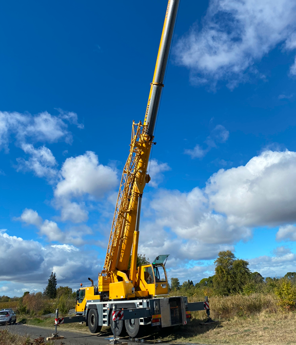 Grue mobile jaune à flèche déployée se détachant sur un ciel bleu nuageux, installée dans une zone herbeuse.