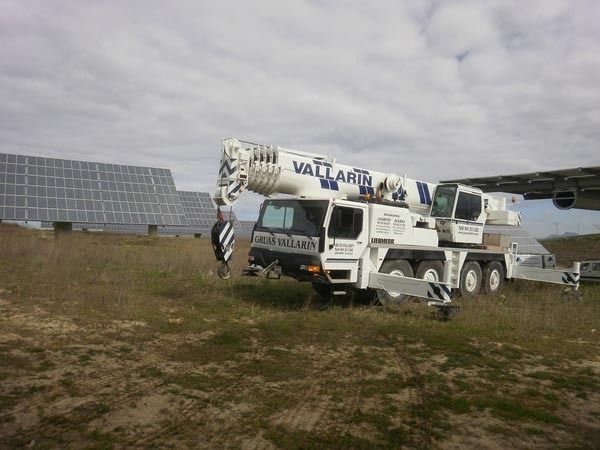 Un camión blanco con una grúa encima está estacionado en un campo con paneles solares al fondo.