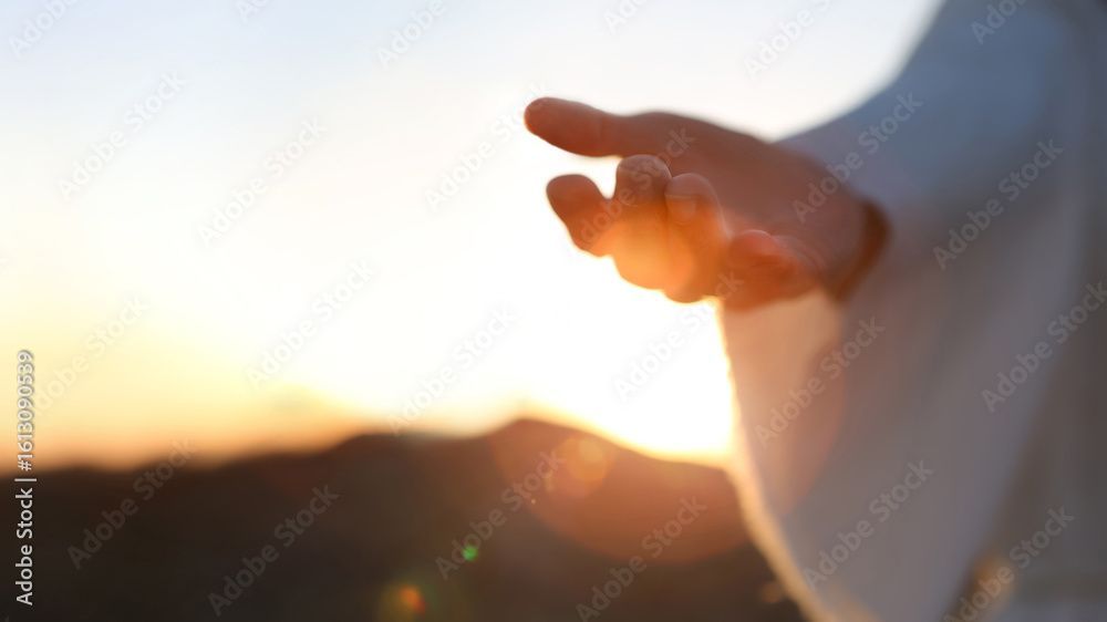 Eine Hand, die dem goldenen Sonnenlicht entgegenstreckt, vor einer verschwommenen Naturlandschaft – möglicherweise ein Gewand.