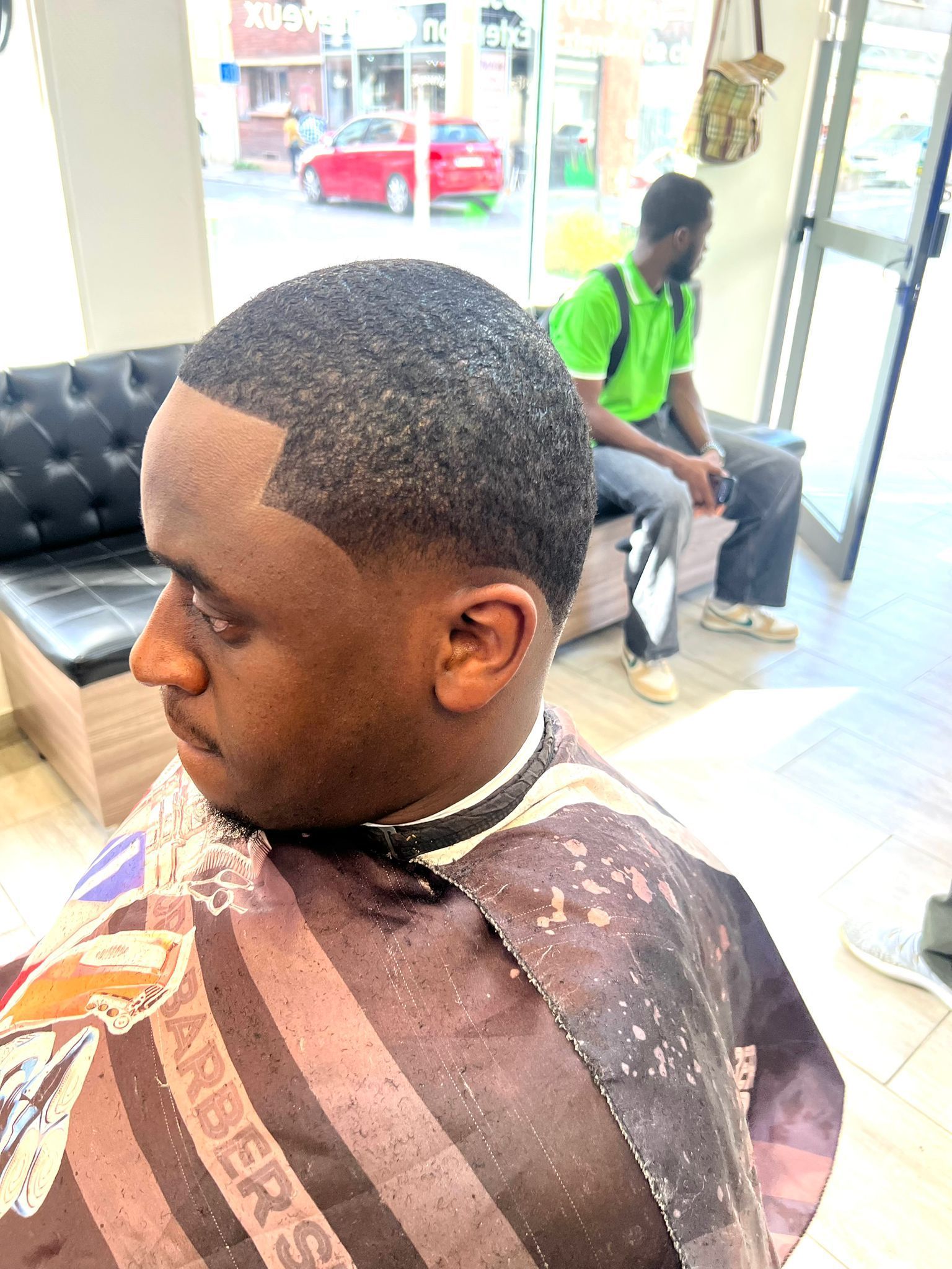 Scène dans un salon de coiffure : un homme se fait couper les cheveux, une autre personne attend sur un canapé près de la fenêtre.