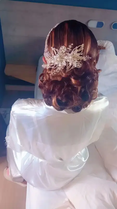 Une mariée en robe blanche, coiffée d'une parure de cheveux ornée, vue de dos, assise à l'intérieur.