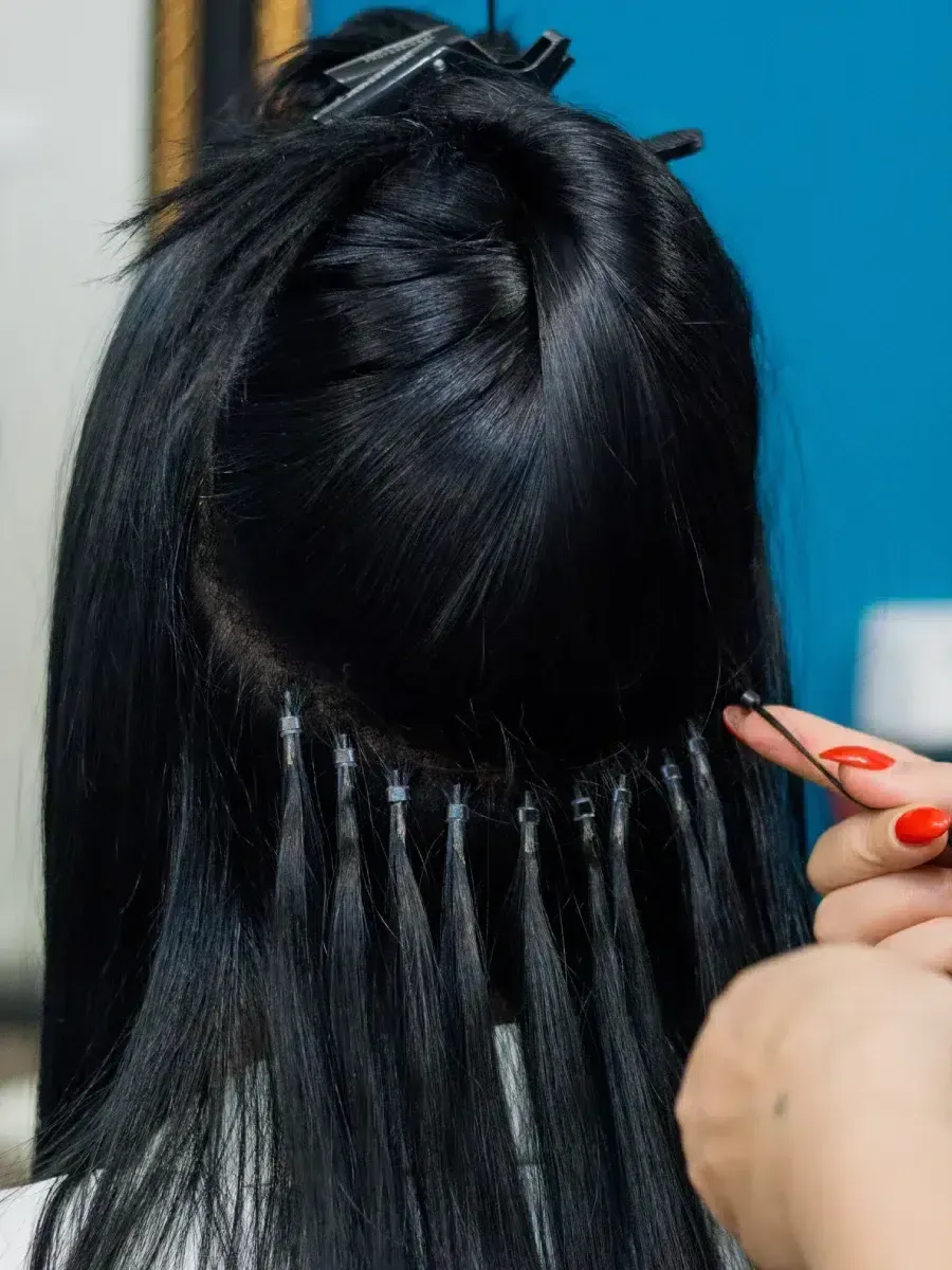 Des extensions de cheveux sont posées sur des cheveux foncés, les mains et les outils étant visibles sur un fond bleu.