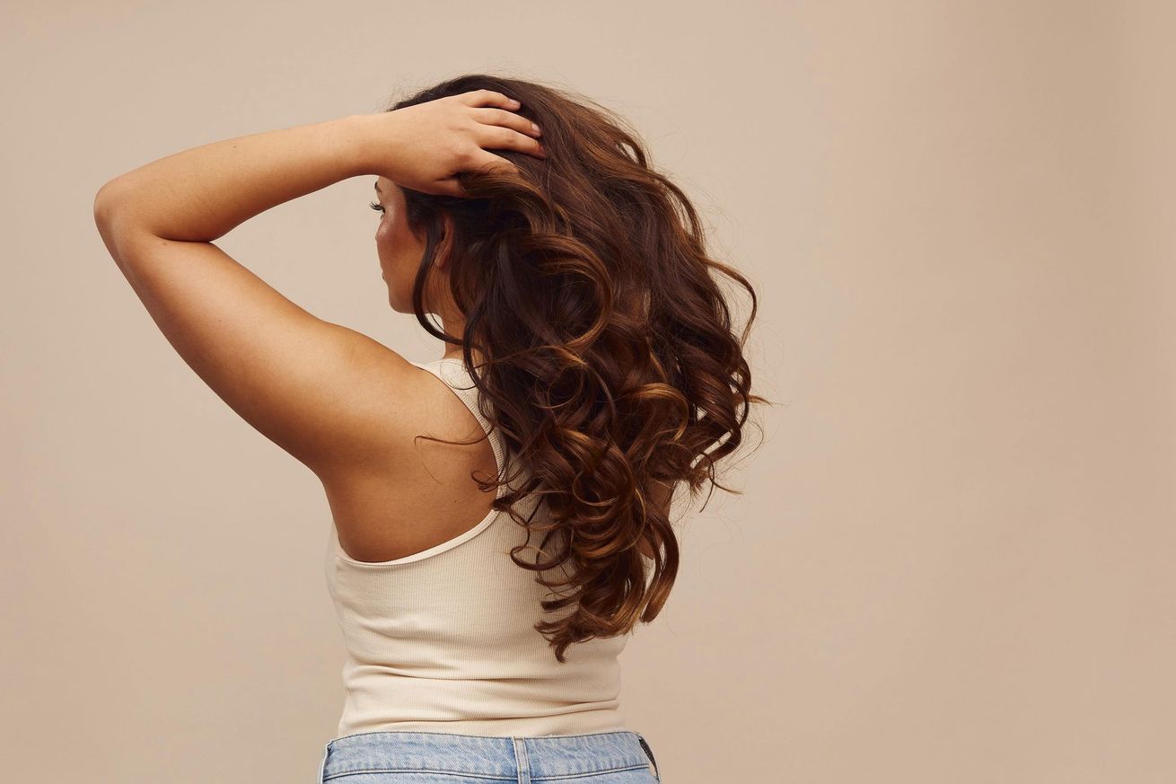 Femme aux longs cheveux ondulés posant sur fond beige, main sur la tête, vêtue d'un haut blanc et d'un jean