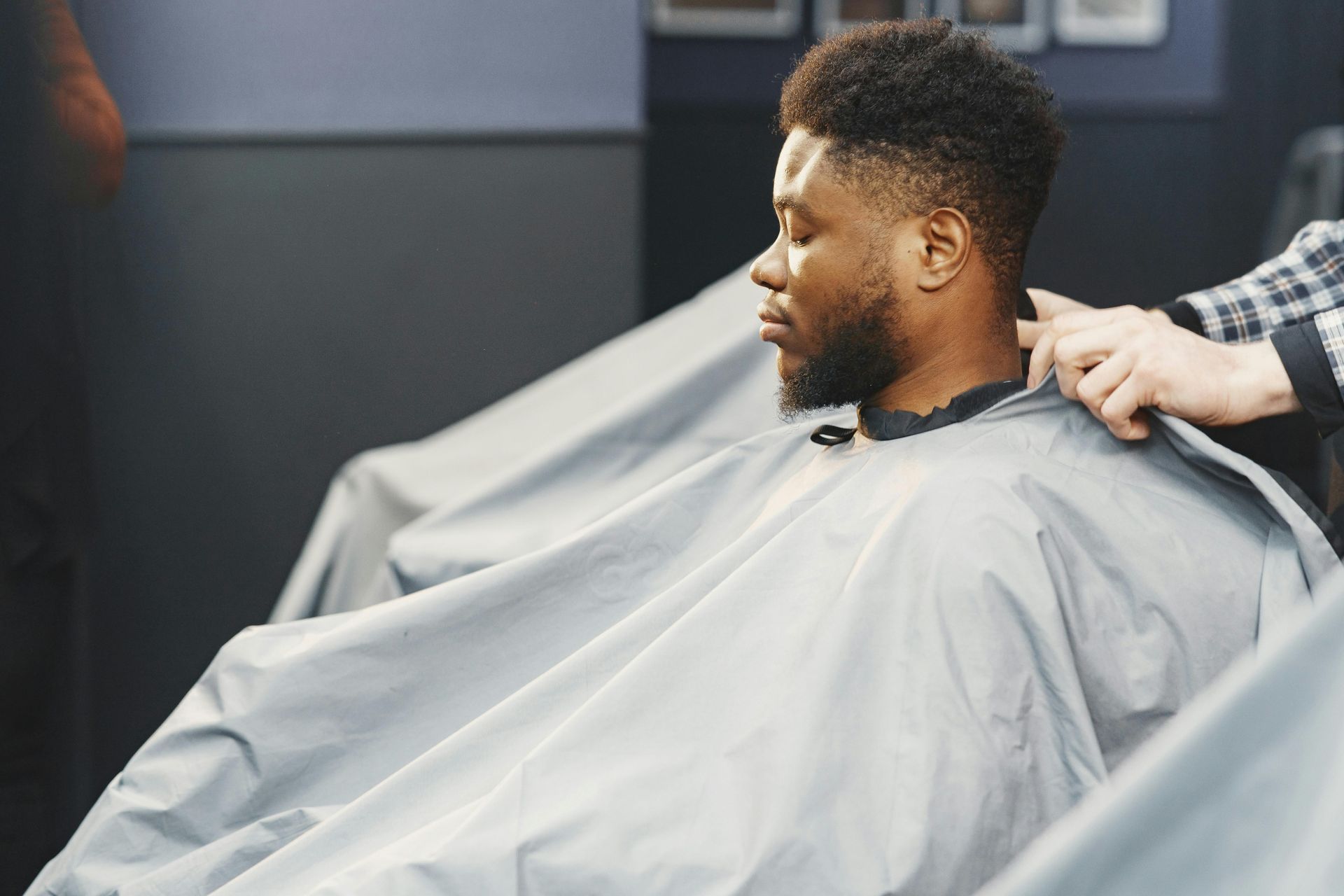 Un homme se fait couper les cheveux dans un fauteuil de barbier, vêtu d'une cape grise, tandis que le barbier lui taille les cheveux.