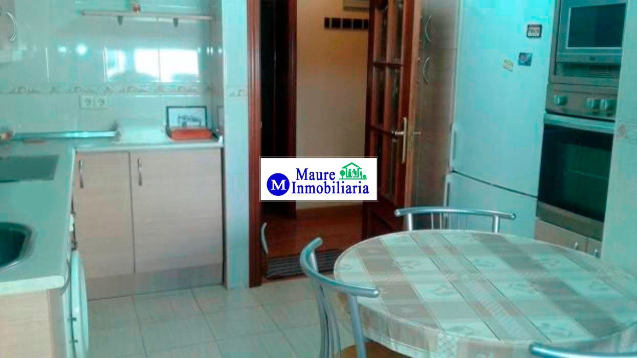 Cocina con armarios, horno, refrigerador y mesa con sillas. La puerta da a otra habitación.