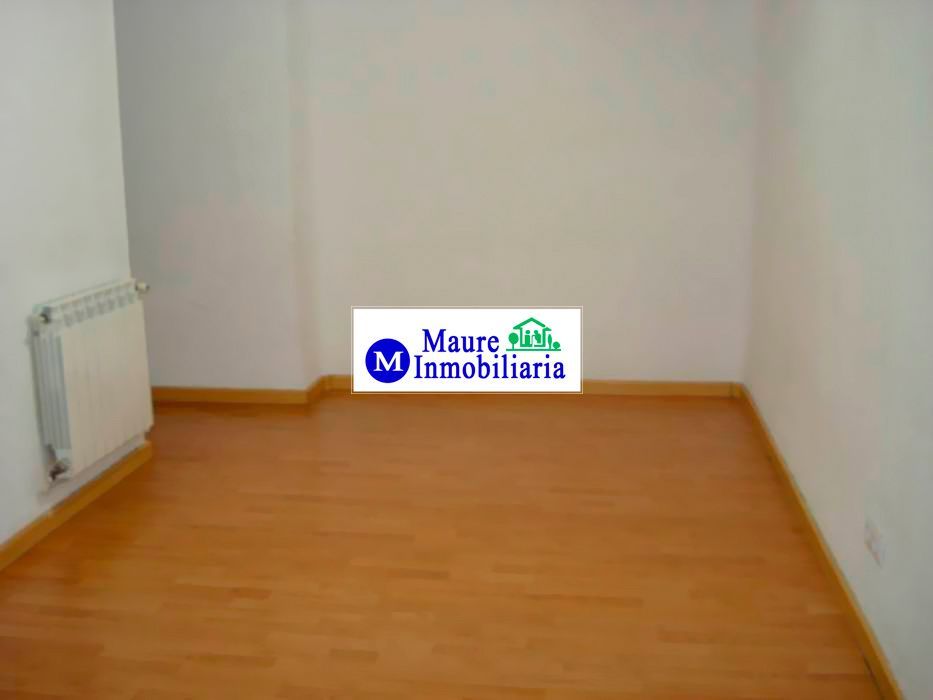 Habitación vacía con suelo de madera, paredes blancas y radiador. Logotipo de una inmobiliaria en la pared.