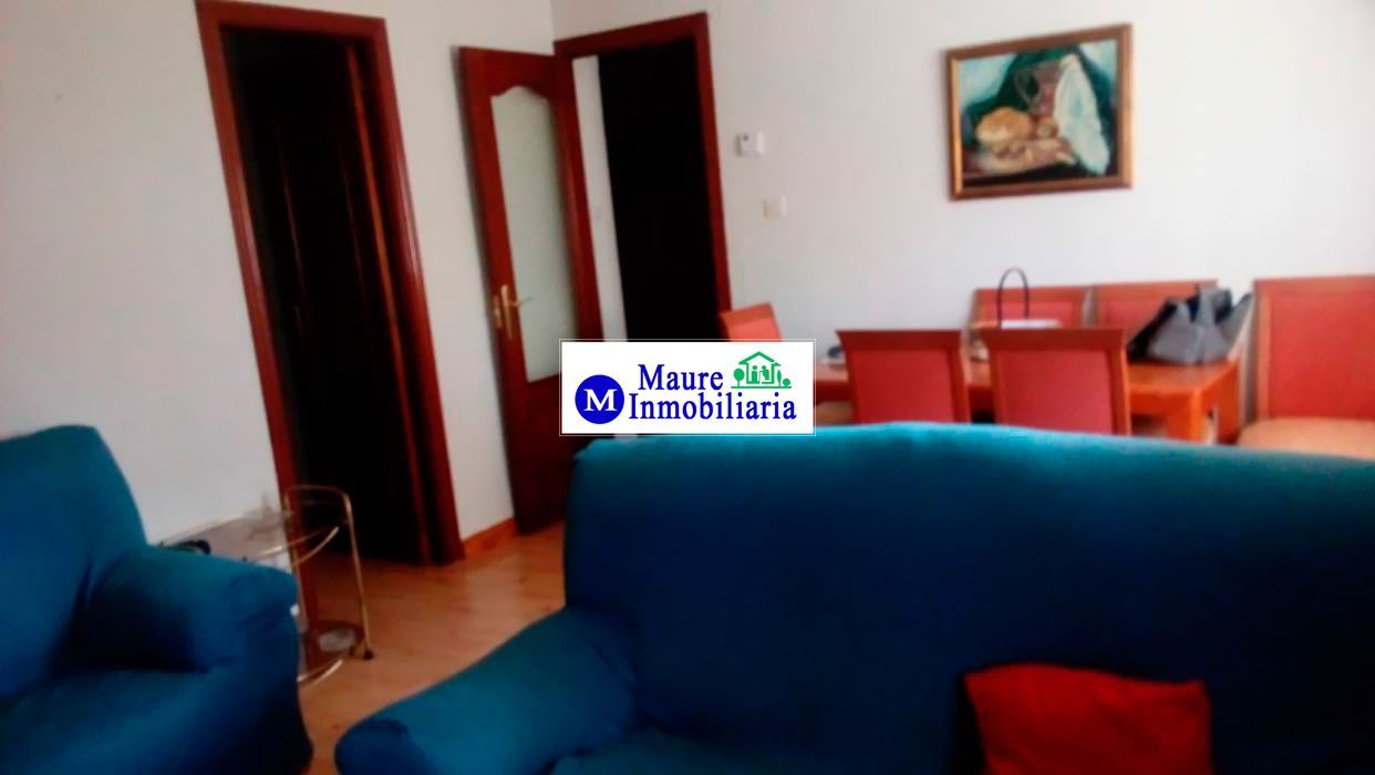 Sala de estar con sofá azul, sillas rojas, mesa de madera y obras de arte. Puertas y suelo marrones.