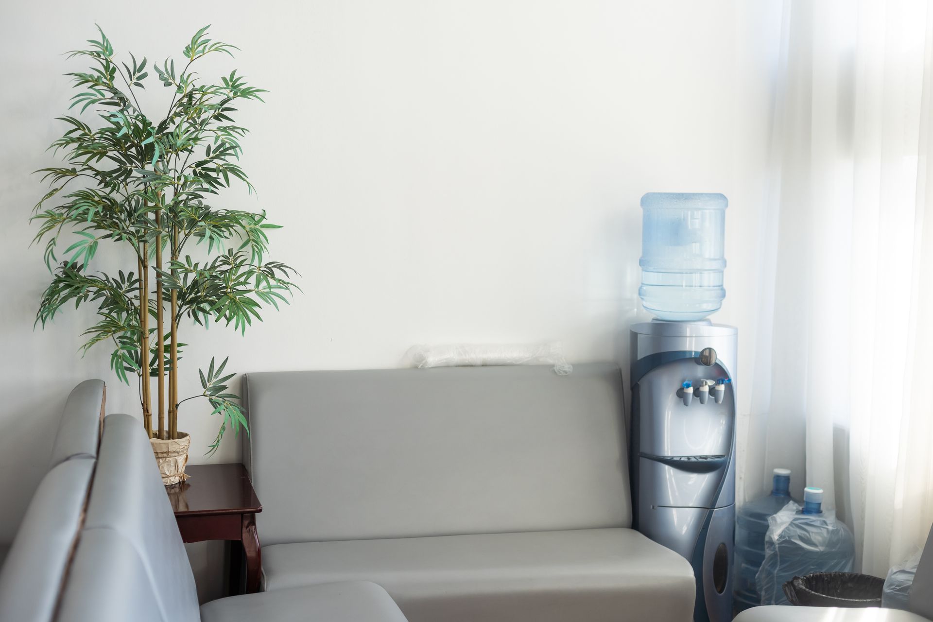 Espace d’attente de bureau avec un canapé gris, une plante en pot et une fontaine à eau près d’une fenêtre