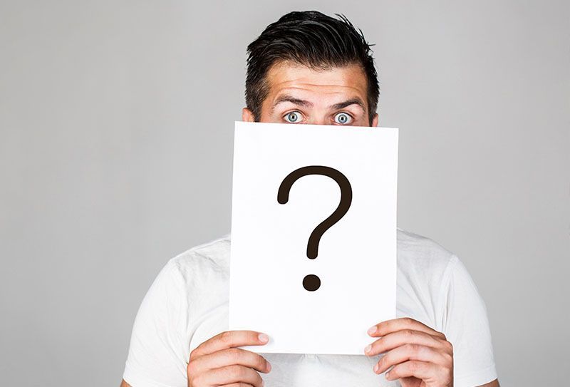 Ein Mann hält ein Blatt Papier mit einem Fragezeichen darauf vor sein Gesicht.