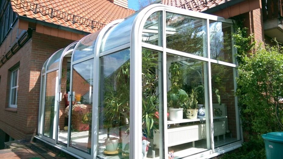 Ein geschwungener, weißgerahmter Glaswintergarten, der an die Seite eines Backsteinhauses angebaut ist und verschiedene Topfpflanzen enthält.
