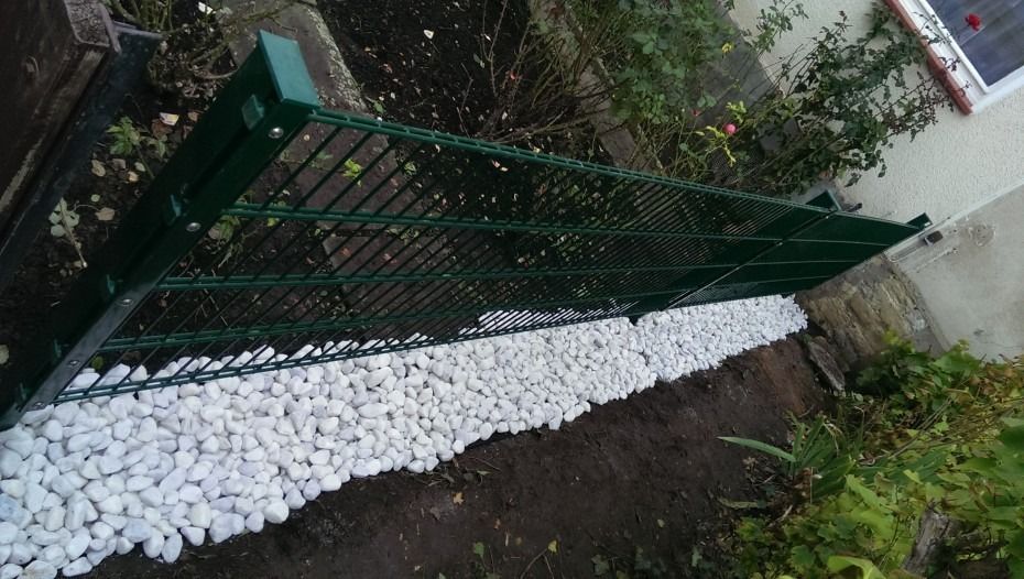 Ein grünes Metallzaunfeld, das über einem mit weißem Zierkies gefüllten Graben am Rand eines Gartens angebracht ist.