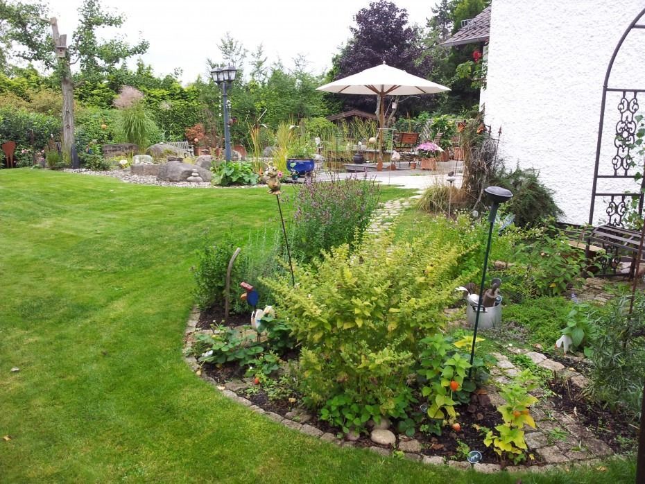 Ein üppiger Garten mit gepflegtem Rasen, einem geschwungenen Blumenbeet mit Sträuchern und einer Terrasse mit weißem Sonnenschirm.