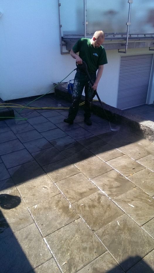 Eine Person in einem dunkelgrünen Hemd reinigt mit einem Hochdruckreiniger eine Steinterrasse vor einem Gebäude.