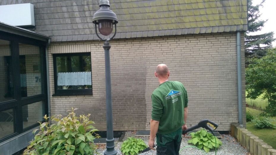 Eine Person in einem grünen Langarmhemd reinigt mit einem Hochdruckreiniger eine verschmutzte Backsteinmauer an der Seite eines Gebäudes.