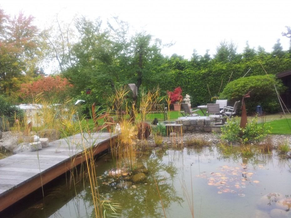 Eine hölzerne Fußgängerbrücke führt über einen Gartenteich vor einer Terrassengarnitur, die von Bäumen und einer dichten Hecke umgeben ist.