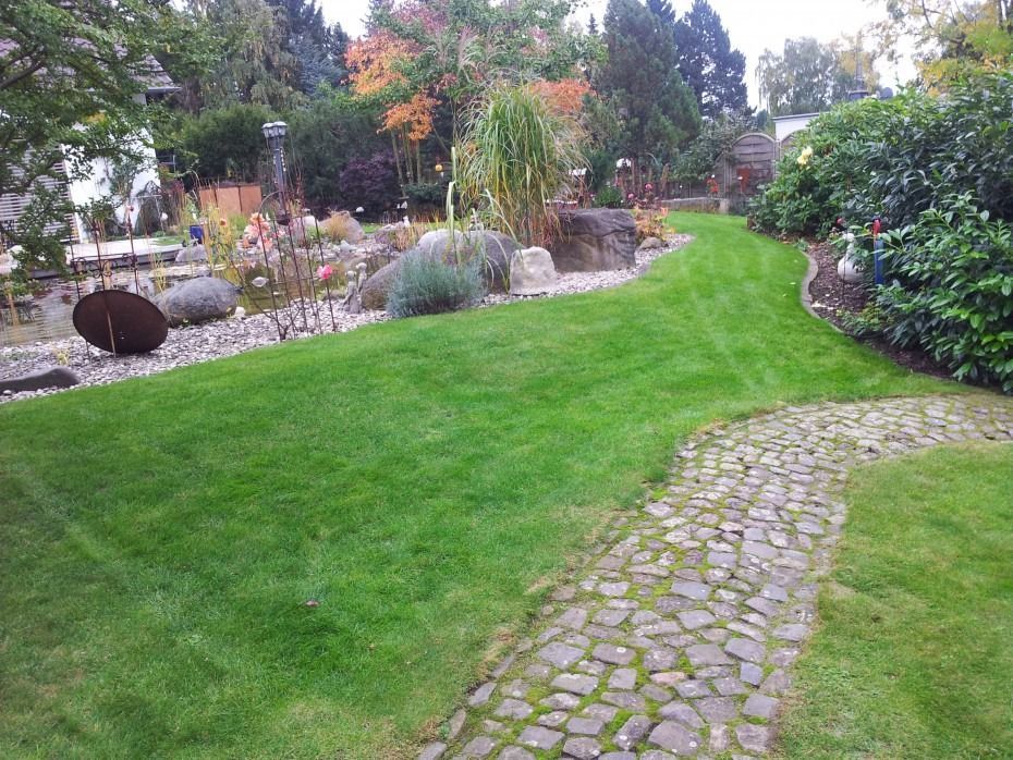 Ein Kopfsteinpflasterweg schlängelt sich durch einen grünen Rasen in einem angelegten Garten mit einem Steingarten und einem Teich im Hintergrund.