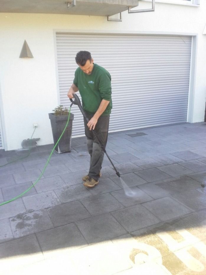 Eine Person in einem grünen Sweatshirt reinigt mit einem Hochdruckreiniger eine dunkel geflieste Terrasse vor einem weißen Gebäude.