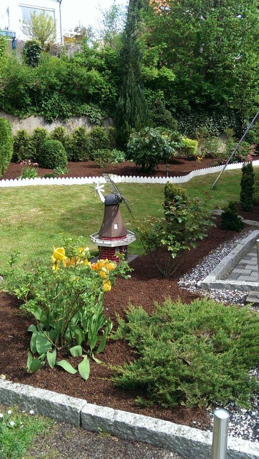 Ein terrassenförmig angelegter Garten mit einer kleinen Windmühle als Ornament, gelben Blumen, Sträuchern und mit Steinen und Zäunen eingefassten Mulchbeeten.